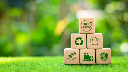 Träklossar med hållbarhetssymboler, som nettonoll, återvinning och minskade CO₂‑utsläpp, staplade på gräs mot en grön, suddig bakgrund. Puukuutioita, joissa on kestävyyssymboleja kuten nettonolla, kierrätys ja CO₂‑päästöjen vähentäminen, pinottuna nurmell
