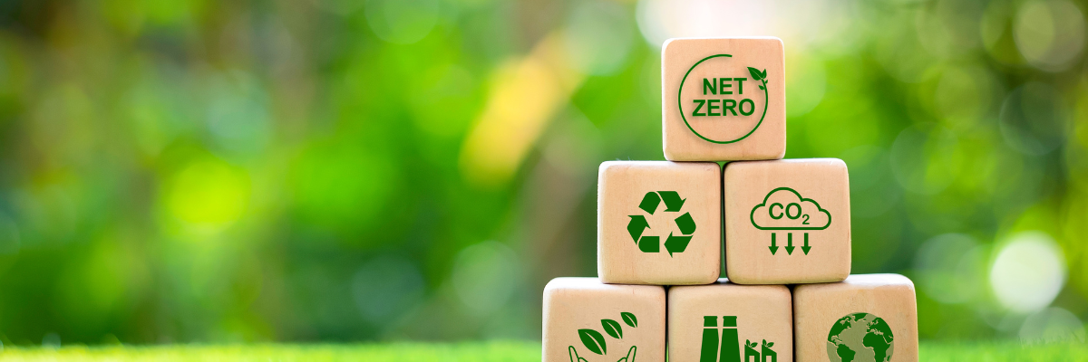 Träklossar med hållbarhetssymboler, som nettonoll, återvinning och minskade CO₂‑utsläpp, staplade på gräs mot en grön, suddig bakgrund. Puukuutioita, joissa on kestävyyssymboleja kuten nettonolla, kierrätys ja CO₂‑päästöjen vähentäminen, pinottuna nurmell