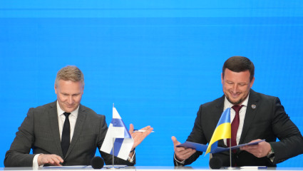 Två personer i kostym sitter vid ett bord och skriver under dokument framför finsk och ukrainsk flagga. Kaksi pukuihin pukeutunutta henkilöä allekirjoittaa asiakirjoja pöydän ääressä Suomen ja Ukrainan lippujen edessä.
