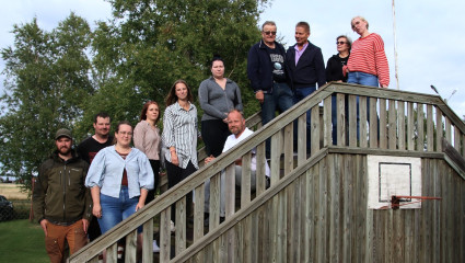 Gruppfoto med elva personer på en trätrappa utomhus, med träd och öppen landsbygd i bakgrunden. Ryhmäkuva yhdestätoista henkilöstä ulkona puisissa portaissa, taustalla puita ja avointa maaseutumaisemaa.