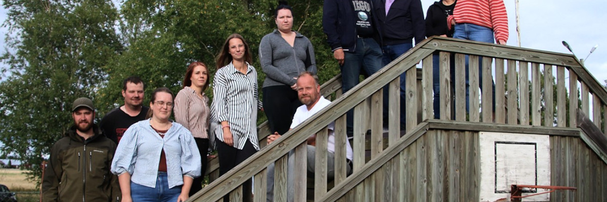 Gruppfoto med elva personer på en trätrappa utomhus, med träd och öppen landsbygd i bakgrunden. Ryhmäkuva yhdestätoista henkilöstä ulkona puisissa portaissa, taustalla puita ja avointa maaseutumaisemaa.