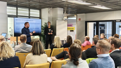 Workshop med publik där två personer presenterar framför en skärm och whiteboard i ett mötesrum. Työpaja, jossa kaksi henkilöä esittelee yleisölle näytön ja fläppitaulun edessä kokoustilassa.