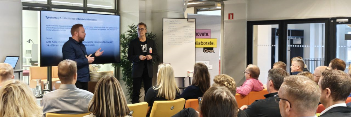 Workshop med publik där två personer presenterar framför en skärm och whiteboard i ett mötesrum. Työpaja, jossa kaksi henkilöä esittelee yleisölle näytön ja fläppitaulun edessä kokoustilassa.