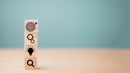 Träklossar staplade på varandra med symboler för sökning, idé, kugghjul och måltavla. Päällekkäin asetetut puupalikat, joissa on hakua, ideaa, rattaita ja maalitaulua kuvaavat symbolit.
