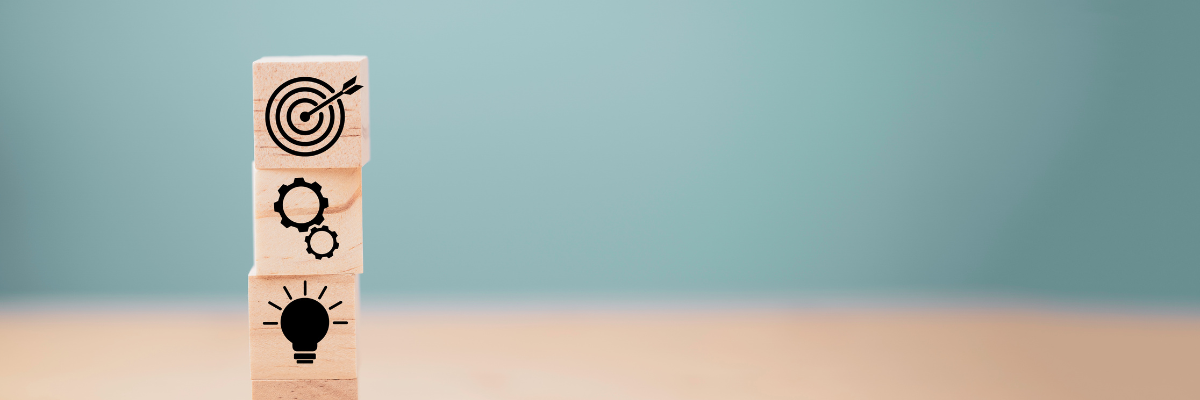 Träklossar staplade på varandra med symboler för sökning, idé, kugghjul och måltavla. Päällekkäin asetetut puupalikat, joissa on hakua, ideaa, rattaita ja maalitaulua kuvaavat symbolit.