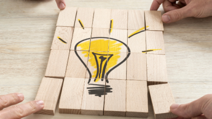 Fyra händer håller träklossar som tillsammans bildar en tecknad gul glödlampa, symbol för idéer och innovation. Neljän käden kokoamat puupalikat muodostavat piirroskuvan keltaisesta hehkulampusta, joka symboloi ideoita ja innovaatioita.