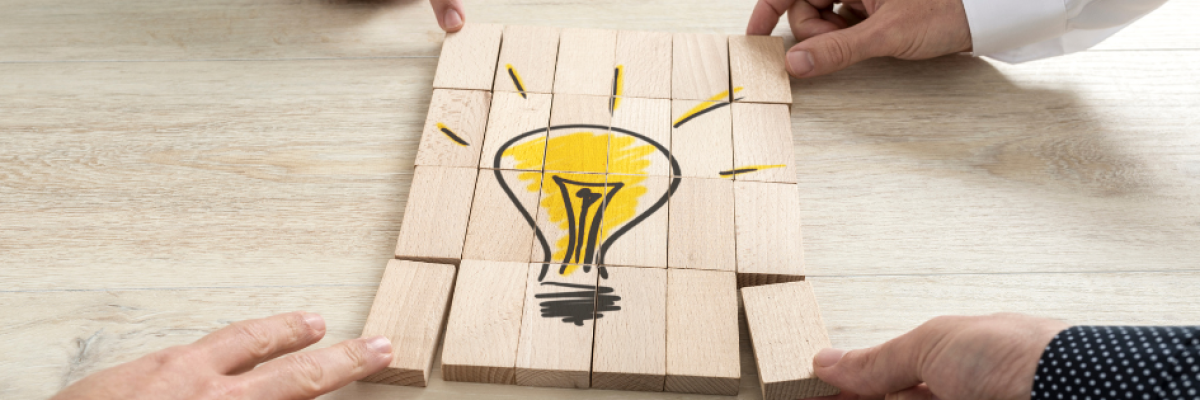 Fyra händer håller träklossar som tillsammans bildar en tecknad gul glödlampa, symbol för idéer och innovation. Neljän käden kokoamat puupalikat muodostavat piirroskuvan keltaisesta hehkulampusta, joka symboloi ideoita ja innovaatioita.